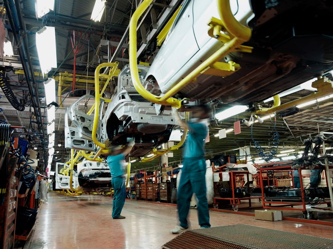 Car factory production line Two workers wearing green shirts and blue pants work underneath a car in an automobile factory. There are assembly lines, partially constructed cars, tools and electrical equipment in the background.